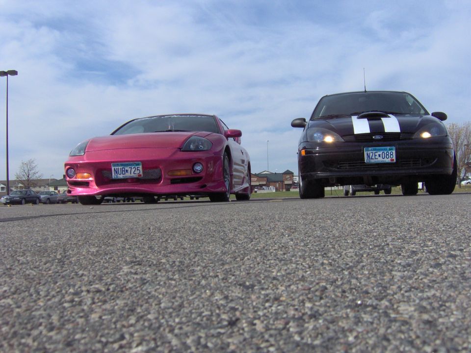 Black Ford Focus with white hood stripes parked beside a pink Mitsubishi Eclipse, low front view