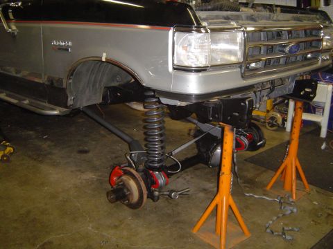 1989 Ford Bronco - The front axle and suspension of a Ford truck are visible as it's supported by jack stands in a garage.