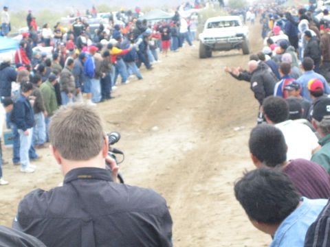 1990 Ford Bronco - white off-road race truck drives down a dusty course as a cameraman and crowd watch from both sides.
