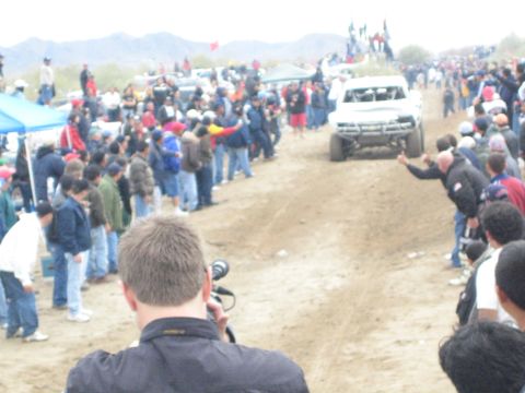 1990 Ford Bronco - white off-road race truck travels a dusty dirt course flanked by a large crowd and desert mountains.