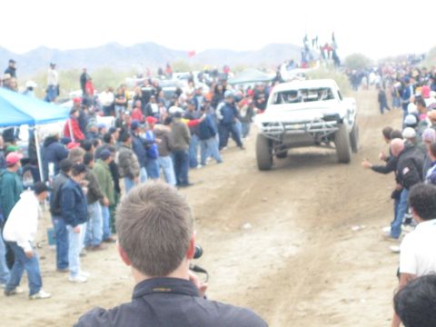 1990 Ford Bronco - white off-road race truck heads down a dirt course as spectators line both sides under a hazy sky.