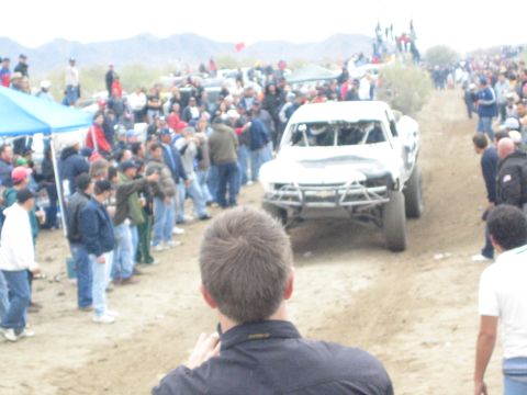 1990 Ford Bronco - white Chevrolet off-road race truck navigates a dirt course as a crowd watches near a blue canopy tent.