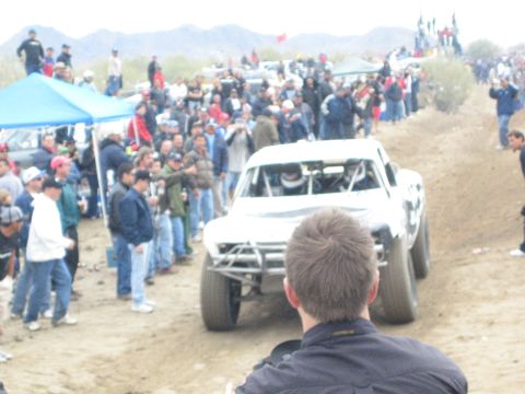 1990 Ford Bronco - white off-road race truck climbs a rutted dirt course as spectators gather near a blue tent in the desert.
