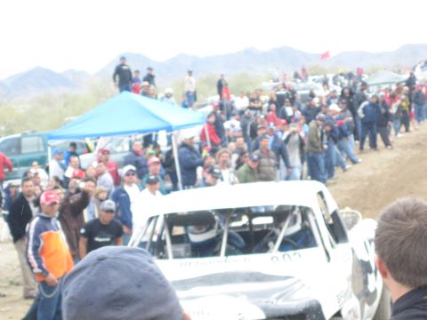 1990 Ford Bronco - white off-road race truck pushes through a dense crowd of spectators on a dirt course in the desert.