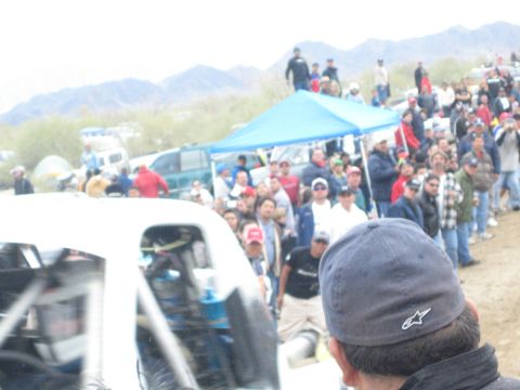 1990 Ford Bronco - man in a gray cap watches as a white off-road race truck idles among a large crowd at a dusty desert event.
