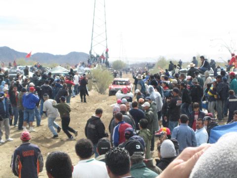 1990 Ford Bronco - Spectators line a dirt track in the desert as an off-road truck races past under a metal tower and power lines.