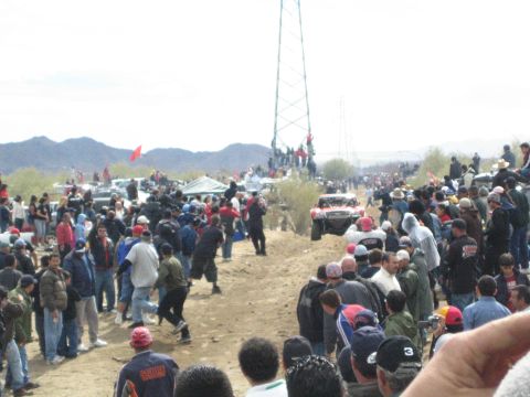 1990 Ford Bronco - large crowd of spectators watches an off-road race truck approach along a dusty desert dirt road.