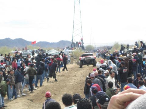 1990 Ford Bronco - off-road race truck speeds down a dirt course flanked by crowds, with desert mountains and a red flag behind.