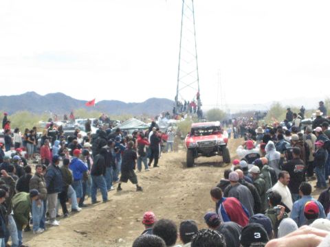 1990 Ford Bronco - red off-road race truck drives between dense crowds on a dirt track at a desert racing event.
