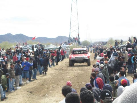 1990 Ford Bronco - red off-road race truck travels a dusty desert track lined with spectators beneath a metal lattice tower.