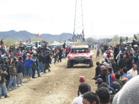 1990 Ford Bronco - red off-road race truck approaches head-on along a dirt course as crowds watch from both sides in the desert.