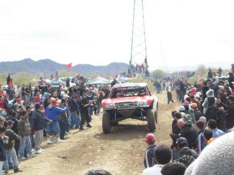 1990 Ford Bronco - red off-road race truck drives toward the camera through a desert crowd lining a dirt track.