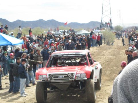 1990 Ford Bronco - red off-road race truck with a Ford grille emblem and Fabtech windshield banner drives a desert dirt course.