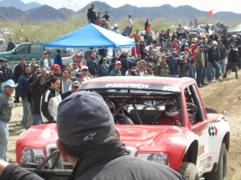 1990 Ford Bronco - red off-road race truck marked Fabtech and number 805 sits among spectators at a desert race under a blue canopy.