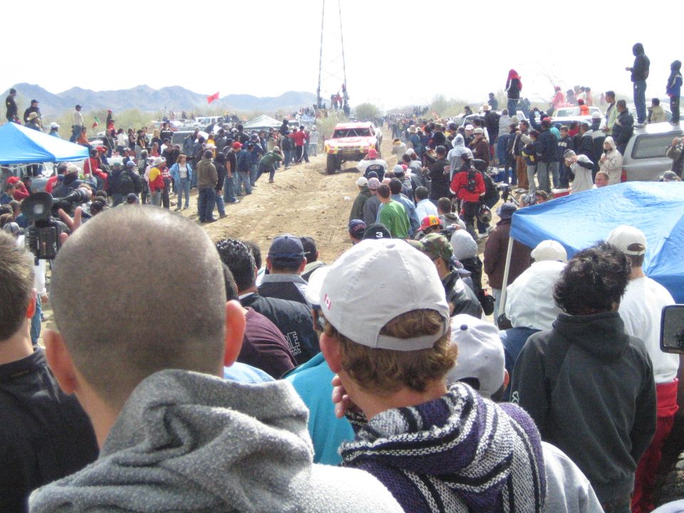 Spectators in caps watching off-road truck on dusty desert course