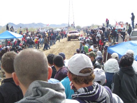1990 Ford Bronco - crowd of spectators watches a distant off-road race truck kicking up dust on a desert dirt track.