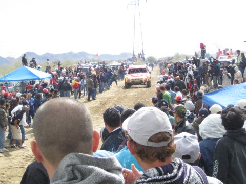 1990 Ford Bronco - white and orange off-road race truck travels a dusty desert track flanked by spectators and blue canopies.