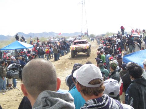 1990 Ford Bronco - off-road race truck kicks up dust along a desert course as spectators and photographers crowd the edges.
