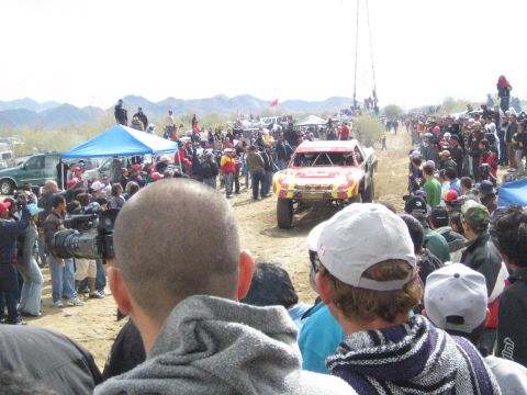 1990 Ford Bronco - red and yellow off-road race truck drives a desert dirt track surrounded by a large crowd of spectators.