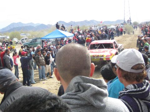 1990 Ford Bronco - red and yellow off-road race truck crests a desert track as spectators watch from a dusty slope.