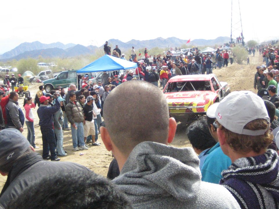 Red and yellow off-road truck cresting desert track near spectators