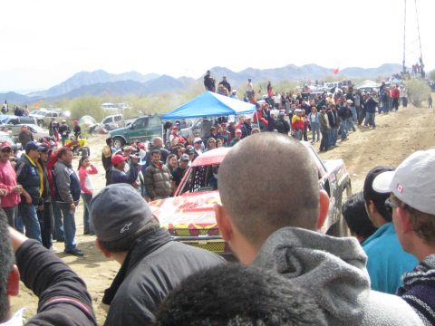 1990 Ford Bronco - red and yellow off-road race truck sits among a dense crowd of spectators at a dusty desert race.
