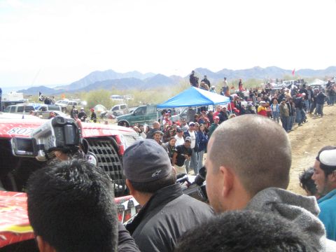 1990 Ford Bronco - red off-road race truck numbered 807 sits among spectators and photographers at a dusty desert racing event.