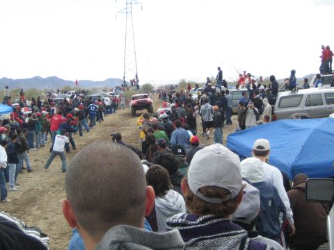 1990 Ford Bronco - off-road race truck approaches down a desert dirt track as spectators and parked trucks crowd both sides.