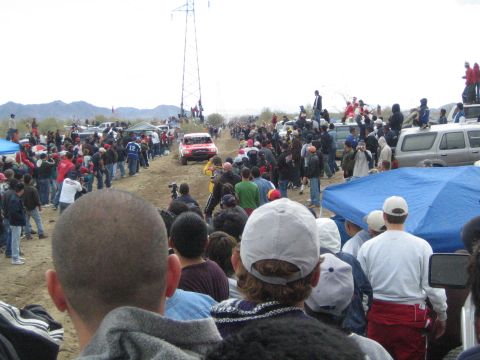 1990 Ford Bronco - Densely packed spectators line a dusty desert course as a red off-road race truck drives toward them under an overcast sky.