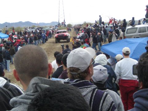 1990 Ford Bronco - red and white off-road truck marked 12 races along a desert dirt road past crowds of standing spectators and parked trucks.