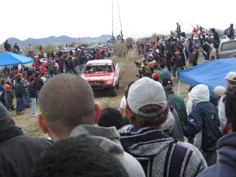 1990 Ford Bronco - red off-road race truck climbs a dirt rise flanked by dense crowds and blue pop-up canopies in a desert setting.