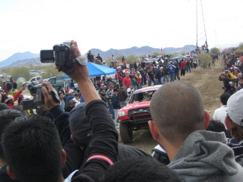 1990 Ford Bronco - spectator raises a camcorder above the crowd while a red Toyota off-road race truck speeds along the dusty desert course.
