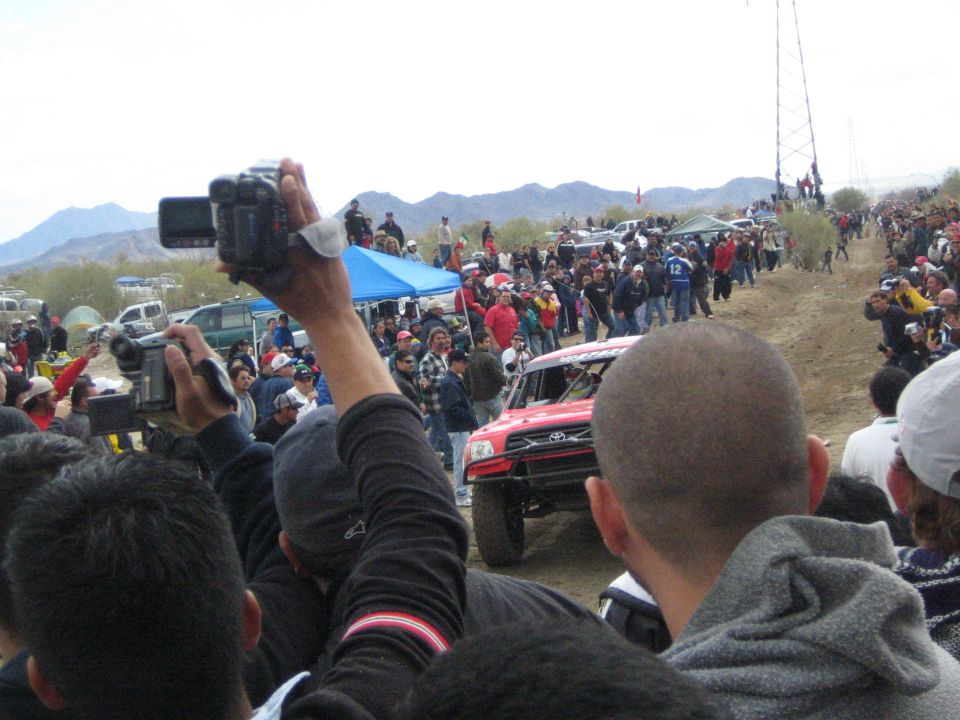 Spectator holds up a video camera as a red Toyota off-road truck races by in the desert