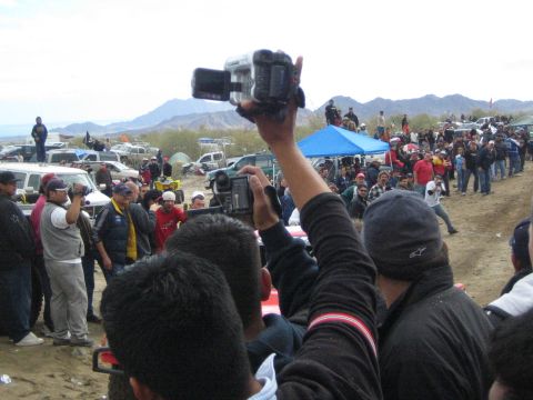1990 Ford Bronco - Fans hold up video cameras over a crowd watching a desert off-road race, with hills and parked vehicles in the background.