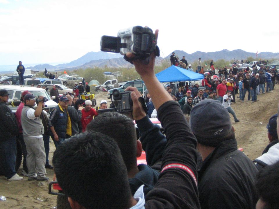 Spectators raise camcorders to film an off-road race in the desert with mountains behind