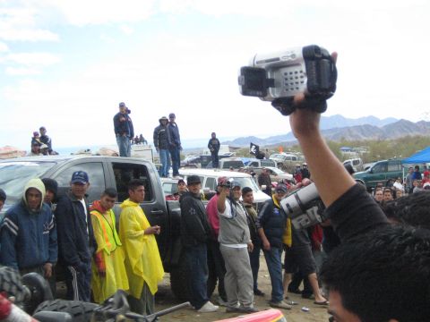 1990 Ford Bronco - Spectators in yellow raincoats stand among parked trucks while a man raises a silver camcorder at a desert off-road event.