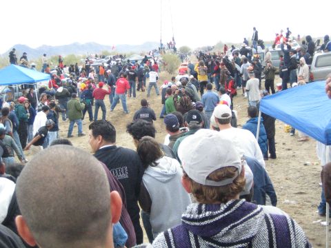 1990 Ford Bronco - large crowd spreads across a dirt desert course with blue tents and parked vehicles as people watch the off-road race.