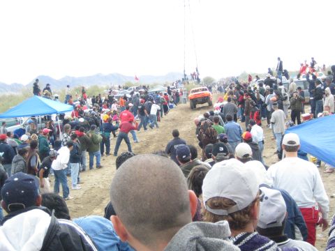 1990 Ford Bronco - orange off-road race truck heads up a dirt rise flanked by spectators and blue canopies in a desert landscape.