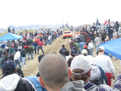 1990 Ford Bronco - orange off-road race truck speeds along a dirt desert course as crowds and a photographer watch from the sidelines.
