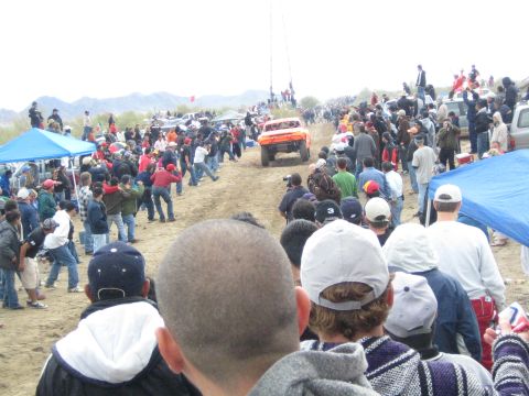 1990 Ford Bronco - orange off-road race truck drives a dirt course while spectators and a kneeling photographer line the desert track.