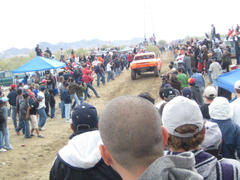 1990 Ford Bronco - orange off-road race truck moves down a dirt desert course tightly lined on both sides by standing spectators.