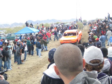 1990 Ford Bronco - orange off-road race truck sits close to a dense crowd on a dirt desert course beside blue canopies and parked vehicles.