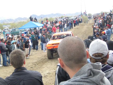 1990 Ford Bronco - orange off-road race truck climbs a dirt slope as crowds of spectators watch from both sides of the desert course.