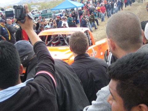 1990 Ford Bronco - orange off-road race truck passes very close to a tight crowd of spectators, one raising a camcorder, in the desert.