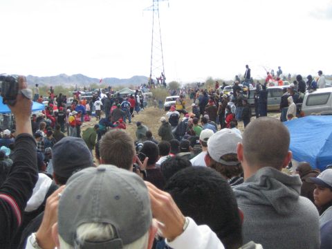 1990 Ford Bronco - Spectators raise cameras to film a desert off-road course where a tall metal tower and parked vehicles stand in the distance.
