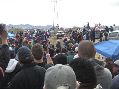1990 Ford Bronco - white off-road truck travels a dirt desert course as a large crowd watches near a tall metal tower under gray skies.