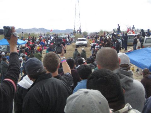 1990 Ford Bronco - Fans hold up video cameras while a white off-road race truck drives a dirt desert course beside a tall metal tower.