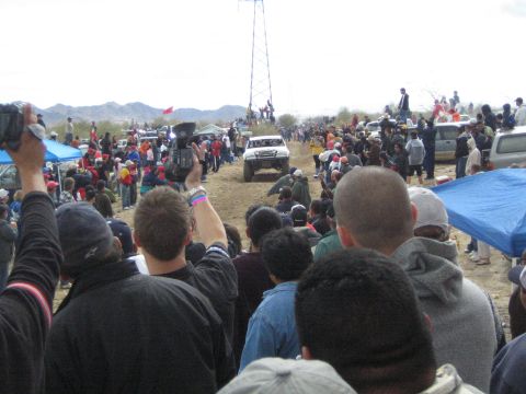 1990 Ford Bronco - white off-road race truck moves along a dirt desert course as crowds film from the sidelines near a tall metal tower.