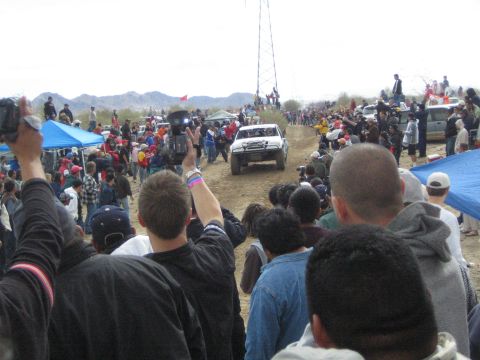 1990 Ford Bronco - white off-road race truck drives a dirt desert course while spectators raise camcorders and watch near a metal tower.