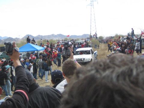 1990 Ford Bronco - white off-road race truck travels a dirt desert course as spectators with cameras and blue tents line the track.